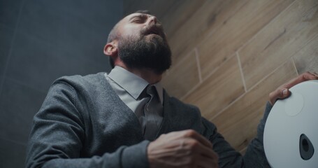 Fototapeta premium Man Sitting on the Toilet and Suffering from Diarrhea and Stomach Ache in Restroom. Businessman Experiencing Discomfort, Indigestion and Digestive Discomfort During Work in Modern Office. Low Angle.