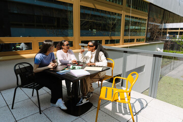 A group of three female students are talking and smiling while sitting at a table with notepads,...