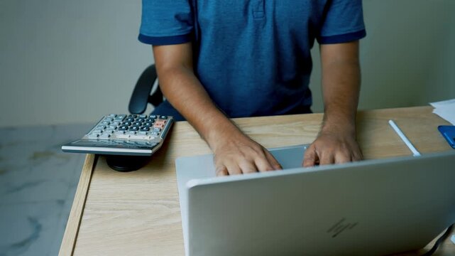 Person working on a laptop at a desk with a calculator and papers