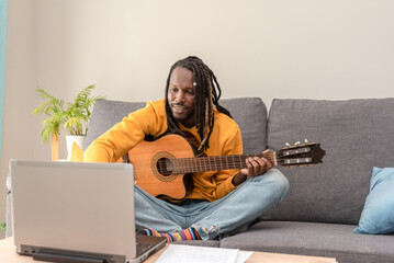 Black man with dreadlocks practicing guitar using online lessons on laptop sitting on a sofa at home