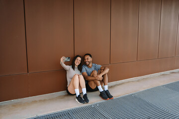 Male and female athletes leaning on a wall and smiling while looking at the phone she is holding...