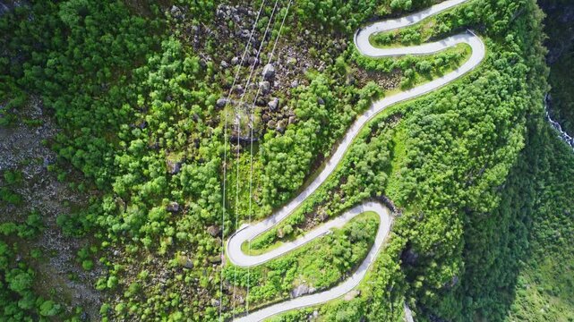 Aerial view of the winding Lysevegen road snaking through lush greenery, adjacent to a cliff and a waterfall, creating a striking contrast of colors, Lysebotn, Rogaland, Norway.