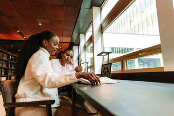 A female student is holding a pen and talking to a female student sitting next to her at a table...