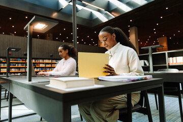 A female student looks into a folder while sitting at a table next to a female student typing on a laptop