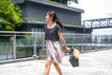 Fototapeta premium Happy asian woman walking outdoors with shopping bags at mall, joyful shopping fashion, consumer happiness, weekend activity and celebrating purchase, sale promotion, cashback reward after shopping