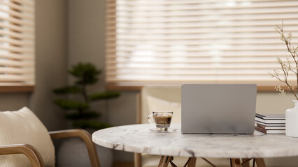 Laptop with coffee cup and books on marble table in a living room with bonsai under window blinds.