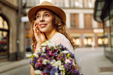 Cheerful woman in hat strolls along sunny street with bouquet of wildflowers. Beautiful woman holds...