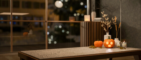 Glowing jack-o-lantern halloween pumpkin and candles on marble table aside glass wall in living room