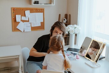A young mother and her 3-year-old daughter play dress-up as makeup artists, sharing laughter and...