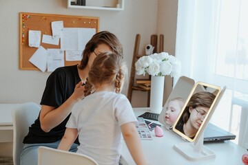 A young mother and her 3-year-old daughter play dress-up as makeup artists, sharing laughter and...