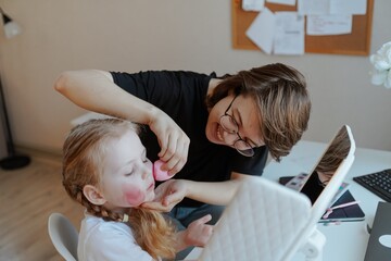A young mother and her 3-year-old daughter play dress-up as makeup artists, sharing laughter and...