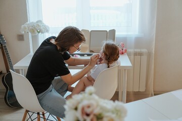 A young mother and her 3-year-old daughter play dress-up as makeup artists, sharing laughter and...