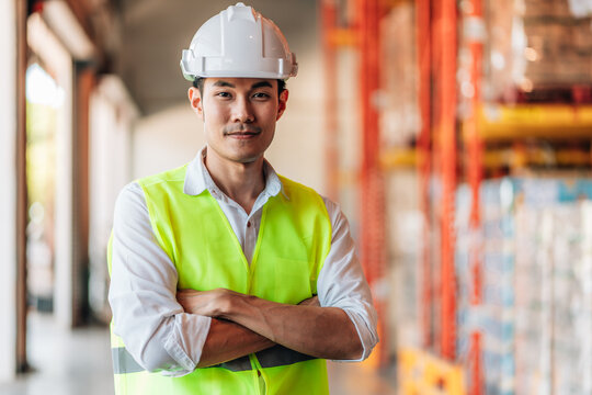 A male warehouse manager in a white shirt, a white hard hat, and a neon green reflective vest stands with his arms crossed. He is looking camera.a sense of professional supervision and order.