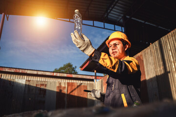 Worker in Safety Gear Holding Plastic Bottle in Recycling Facility Under Bright Sunlight with Warehouse Background