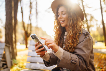 Portrait of cheerful woman in hat, using phone, sitting on bench in autumn park. Beautiful woman...