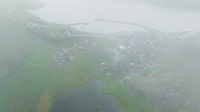 Aerial view of the village of Ei&eth;i, with its harbor and surrounding green landscape, appearing serene under a blanket of mist, Ei&eth;i, Eysturoy, Faroe Islands.