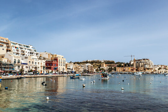 Panoramic view of the Marsaskala waterfront with colorful buildings, boats, and clear turquoise water on a sunny spring day, Malta, Europe