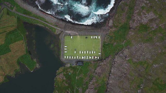 Aerial view of Ei&eth;i Camping's vibrant green football pitch, contrasting sharply with the rugged coastline and crashing waves nearby, Ei&eth;i, Eysturoy, Faroe Islands.