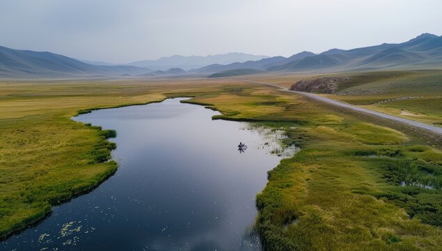 Serene aerial view of a tranquil lake nestled within a vast, flat expanse of grassland, mountains in the distance under a muted sky. A small boat is visible on the lake