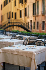 Empty outdoor restaurant tables with tablecloths and cutlery in Venice, Italy. Street cafe with bridge over Venetian canal on background in morning. Concept of dining, tourism and travel