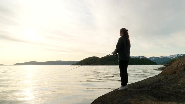 Woman casts pole and fishes in Tromso fjord under soft light, surrounded by still water and hills