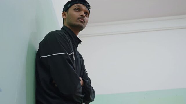 Young man wearing black jacket and headband leaning against wall with arms folded, thoughtful and calm expression captured from low angle, highlighting presence, posture, and concentration