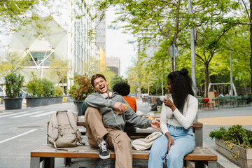 A male student laughs and holds a pastry while sitting on a bench next to a female student holding a cup