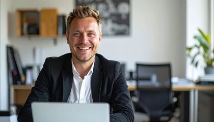 a smiling thirty blonde man with short hair wearing a black jacket and a white shirt sitting at the desk in the office