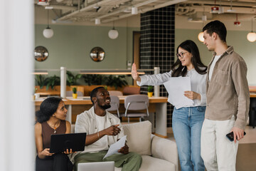 A female worker holds her hand in front of her while holding papers while a male worker stands next to her and a male and female workers sit on the couch
