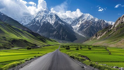 A scenic road winds through a valley, flanked by vibrant green fields and snow-capped mountains under a partly cloudy sky
