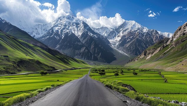 A scenic road winds through a valley, flanked by vibrant green fields and snow-capped mountains under a partly cloudy sky
