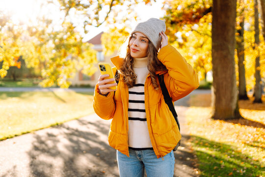 A young woman in a bright jacket with a phone in an autumn forest. A beautiful woman enjoys the sunny weather and nature with her phone outdoors. Concept of nature, technology.
