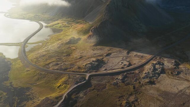 Aerial view of a winding road near a body of water, with mountains in the background, showcasing the rugged landscape, Hvalnes Lighthouse, Sveitarf&eacute;lagi&eth; Hornafj&ouml;r&eth;ur, Iceland.
