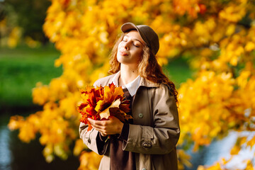 A young woman in a raincoat stands with a bouquet of fallen leaves in an autumn park. A beautiful tourist strolls and enjoys the sunny weather outdoors. Concepts of fashion, seasonality, and leisure.