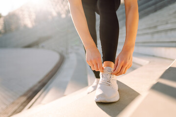 Close-up: A young athlete in sneakers ties her shoelaces on a sports field. A woman in athletic shoes trains outdoors. Sports and training concept. Active lifestyle.