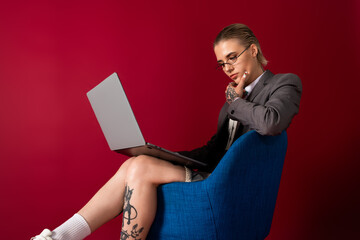 Stylish young woman sitting in a blue chair while working on a laptop against a red background
