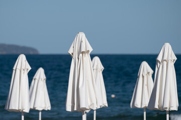 Rows of closed white beach umbrellas stand neatly on a sunlit shore, facing a calm blue sea under a clear sky.