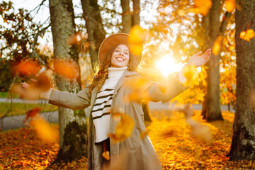 A happy woman is having fun in an autumn park at sunset. A beautiful woman in casual clothes throws leaves into the air on a sunny day. Concept of a walk, relaxation.