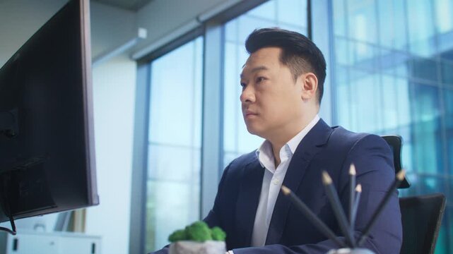 Focused Asian businessman in formal navy suit typing at desk in daylight office. Entering project notes or preparing contract draft. Concentrating on task or finalizing written proposal.
