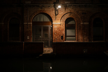 Fototapeta premium Venetian facade at night with glowing lamplight highlighting textured red brick, arched windows, and a wooden doorway above a canal, evoking history and quiet urban atmosphere