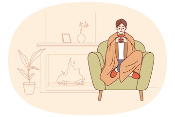 Man feels comfort and harmony sitting by fireplace with blanket on shoulders and cup of hot coffee in hands. Guy enjoys comforts of home and apartment is cozy, allowing him to relax on winter evenings