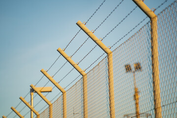 Tall barbed wire fence and security lighting structure under blue sky, symbolizing guarded...