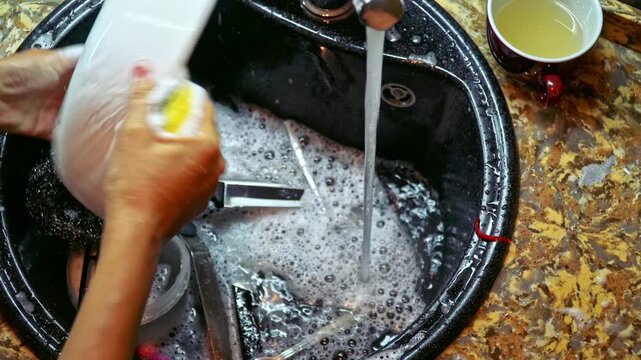Top view woman washing dishes at kitchen sink and placing them on drying rack, focus on hands and tableware, authentic domestic cleaning 4K video footage.