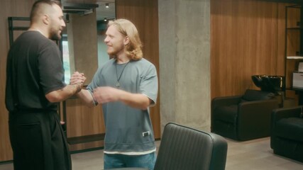 Medium shot of stylish young Caucasian barber with tattoos sitting in chair, browsing online on smartphone, greeting and hugging regular customer arriving for hair cutting and grooming appointment