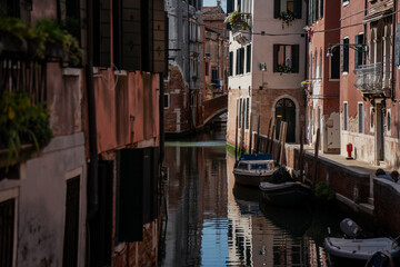 Narrow Venetian canal with arched pedestrian bridge, moored boats and weathered facades reflecting...