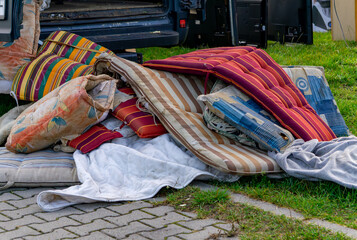 Flea market of antiques with used mattresses, pillows, and colorful garden cushions placed on the ground beside a car