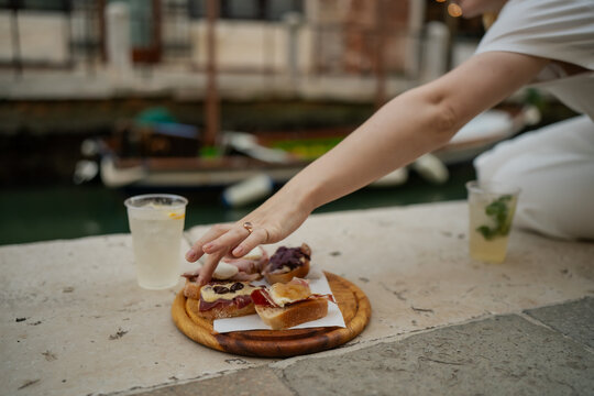 Close-up of hand reaching for Venetian cicchetti selection with cured meats, cheeses and spreads served on bread with spritz cocktails by canal in Venice Italy