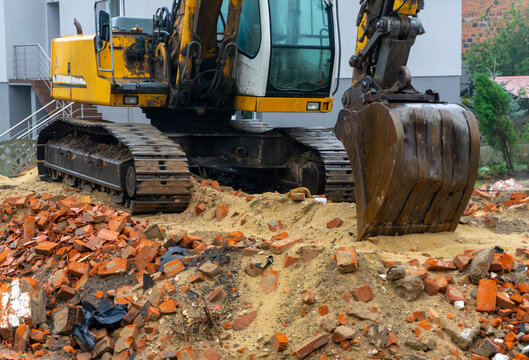 Brick rubble from building demolition with standing tracked excavator nearby, bucket resting in sand at active construction site.