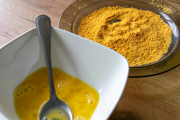 Bowl with beaten egg and spoon next to plate with yellow breadcrumbs, preparation of ingredients for frying dish.