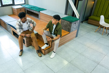 Two Young Male Friends Chatting Casually While Sitting on Bench in Modern Office Lounge